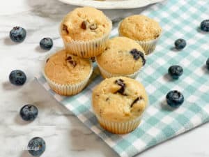5 muffins on a blue checkered cloth with blueberries scattered