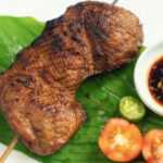 A grilled veggie chicken inasal on a banana leaf, served with sliced tomatoes and dipping sauce.