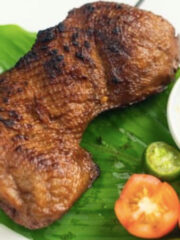 A grilled veggie chicken inasal on a banana leaf, served with sliced tomatoes and dipping sauce.