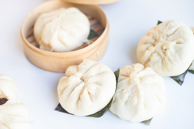 steamed vegan siopao in a bamboo steamer and some on a white surface with bamboo leaves at their bottom