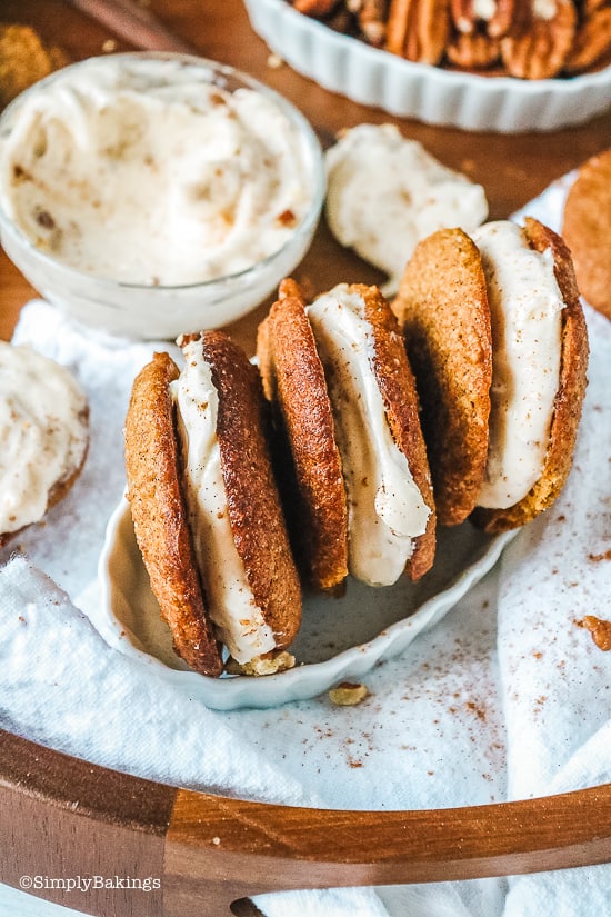3 pumpkin whoopie pies in a white dish surrounded by a bowl of cream cheese and pecans 