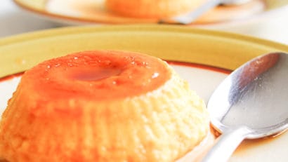 Close-up of a leche flan topped with a bright orange, jello-like layer on a yellow plate, with a spoon beside it.