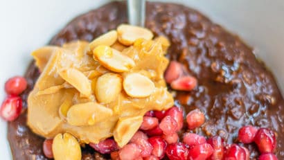 a bowl of chocolate oatmeal with a stainless steel serving spoon and topped with pomegranate seeds, peanut butter and peanuts