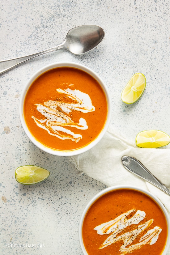 two bowls of cream of tomato soup beside a stainless spoon and slices of lime