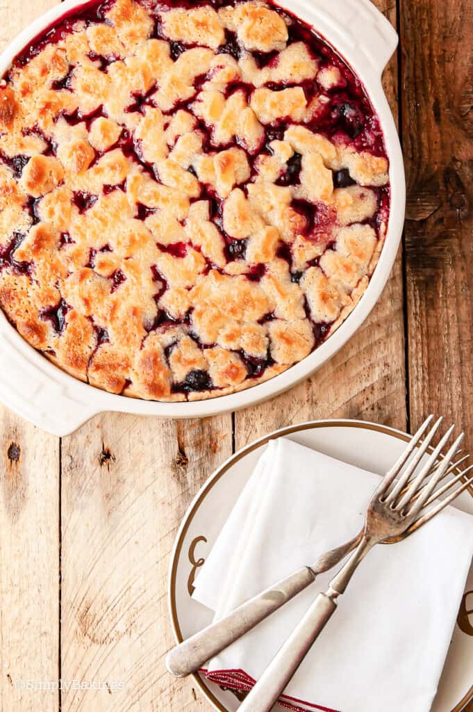 mixed berry cobbler served in a white bowl beside a a dessert plate with a pair of stainless spoon and fork