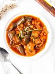 a bowl of hearty eggplant chickpea stew beside a stainless spoon and pile of books