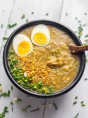 tasty vegetarian congee in a black bowl with ladle topped with boiled egg, green onions and toasted garlic