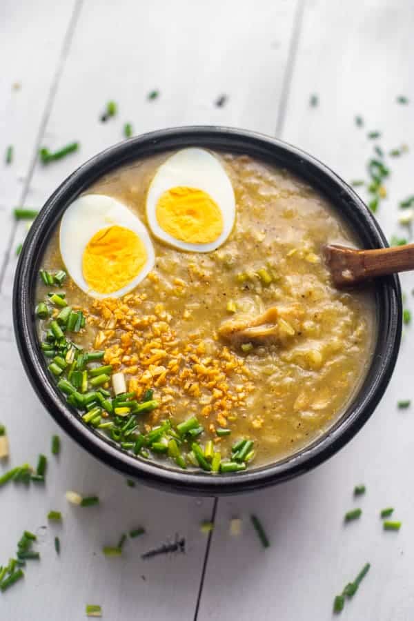 tasty vegetarian congee in a black bowl with ladle topped with boiled egg, green onions and toasted garlic
