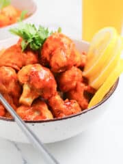 a bowl of vegan cauliflower bites with huli huli sauce, garnished with cilantro and lemon slices with fork on the side, image created by SimplyBakings