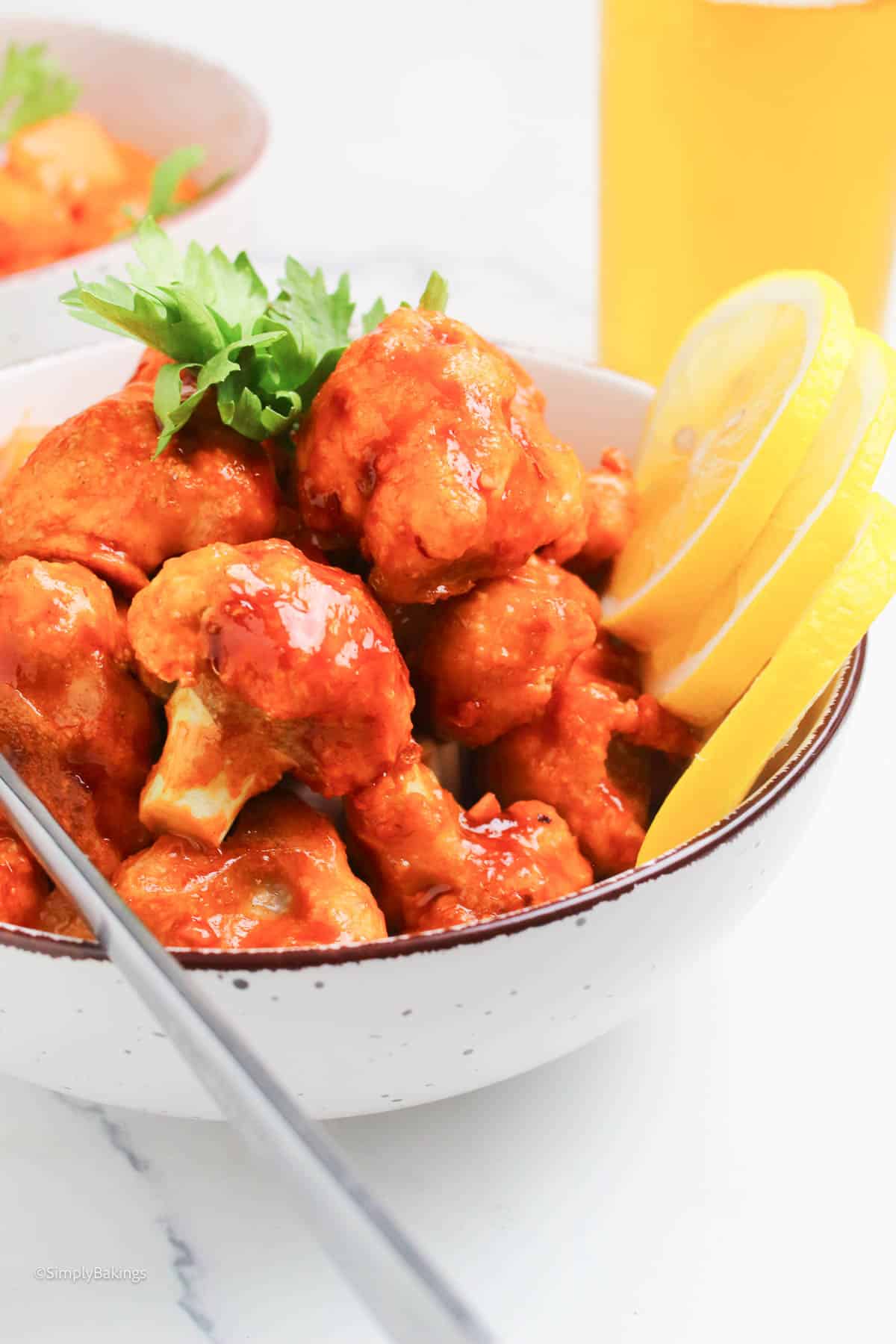 a bowl of vegan cauliflower bites with huli huli sauce, garnished with cilantro and lemon slices with fork on the side, image created by SimplyBakings