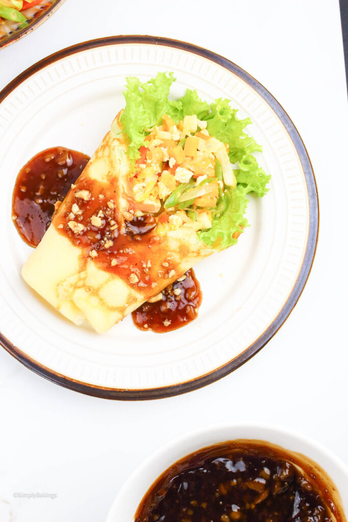 a small bowl of peanut sauce and a lumpiang sariwa served on a white plate drizzled with a peanut sauce