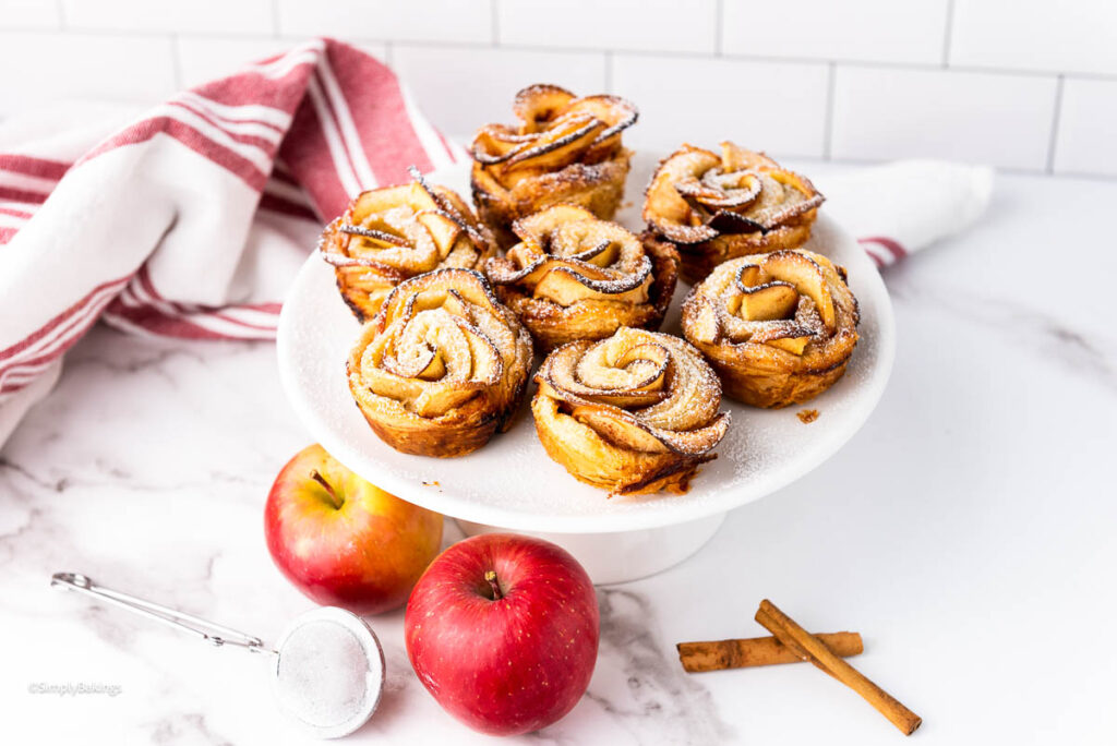 Apple Roses dusted with powdered sugar served on a white dessert stand beside two apples, cinnamon sticks and strainer