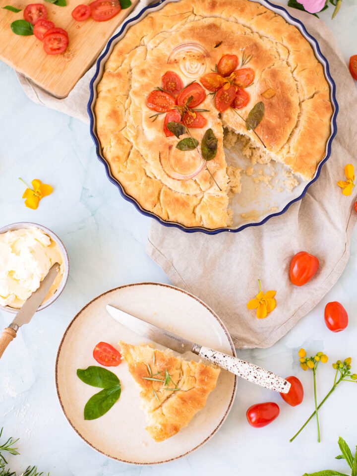 freshly baked Focaccia with Tomatoes and Onions on a circular pan and a slice of it on a small white plate with a knife
